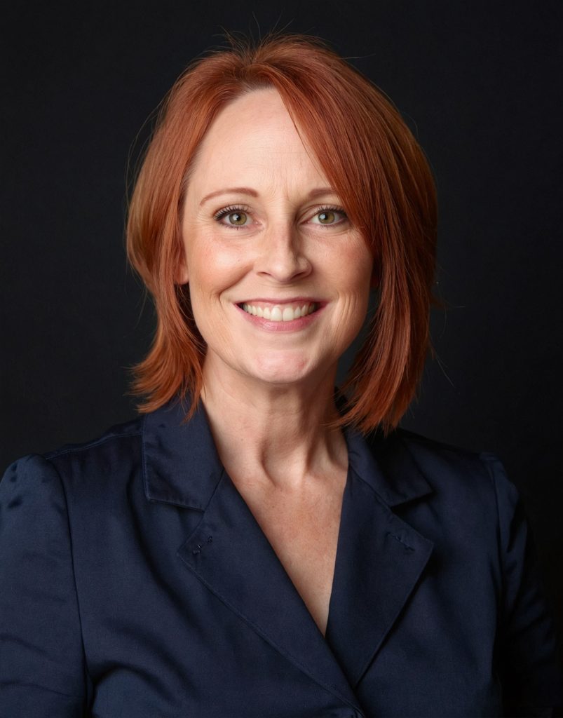Headshot of Bobbie. She has red, shoulder-length hair, green eyes and freckles and is smiling directly at the camera. She's wearing a dark blue suit on a black photo background.