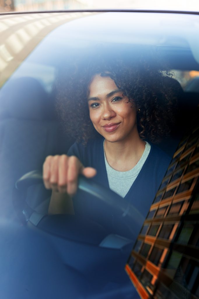 Professionally-dressed woman with brown skin and shoulder length curly hair, smiling while driving in downtown, with a reflection of a downtown high rise in the windshield. 