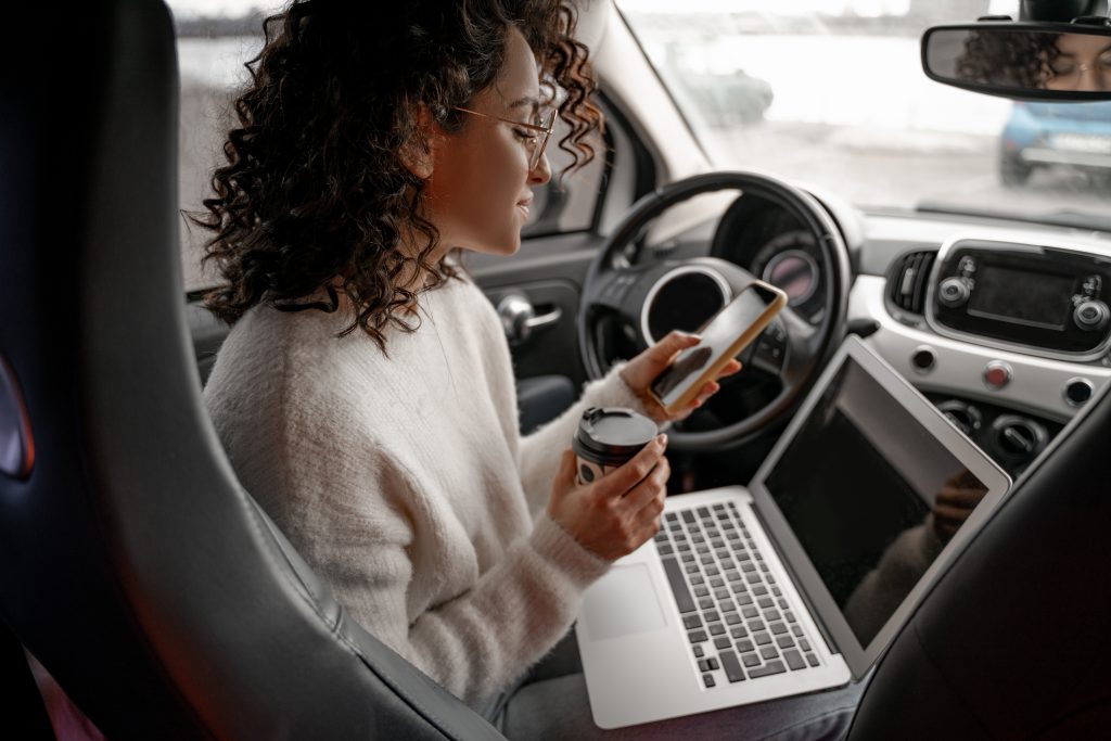 Woman sitting in driver's seat of parked car. She has brown skin, round glasses with gold metal rim, brown curly hair. She is hold a small coffee and her smartphone with her open laptop on her lap.
