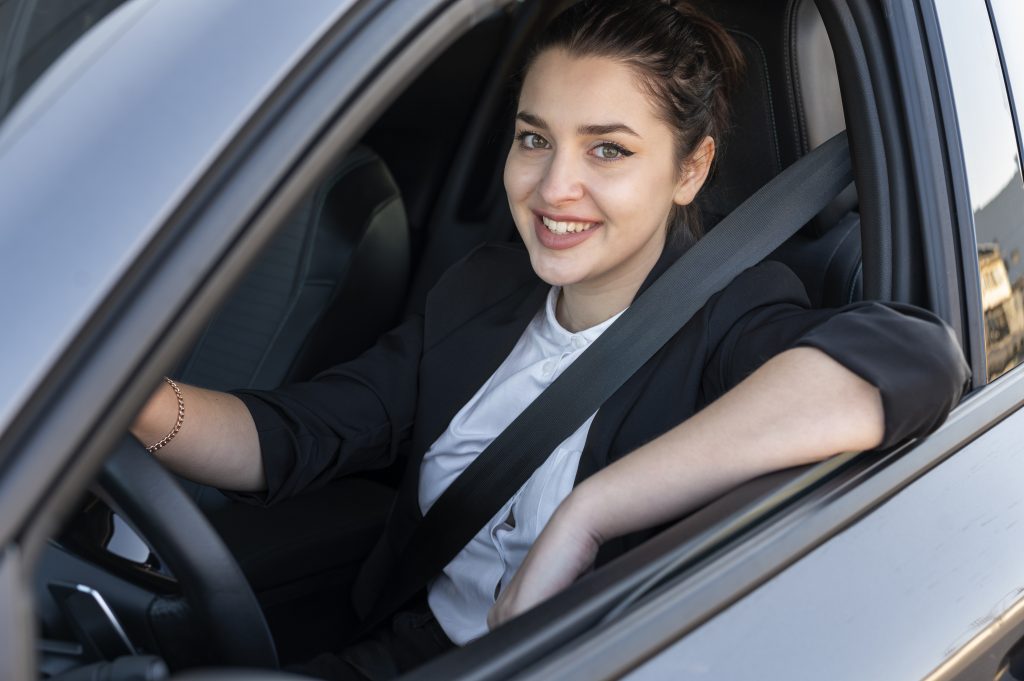 Young female driver with seatbelt on, smiling at the camera, window down, with her arm up on the edge of the door.